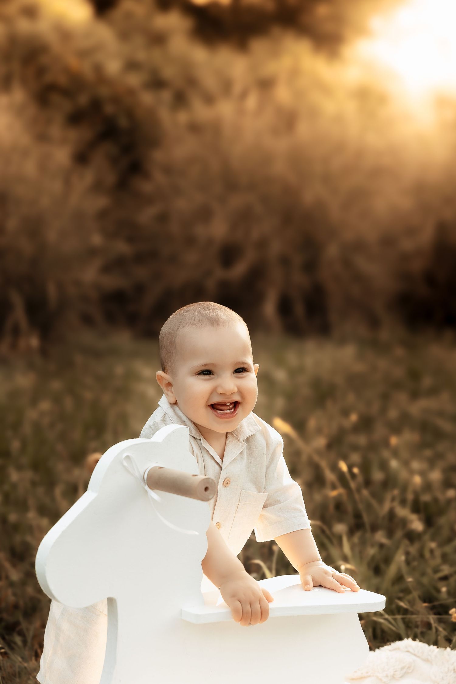 Smiling baby in white outfit playing with a white rocking horse in a golden sunset field.