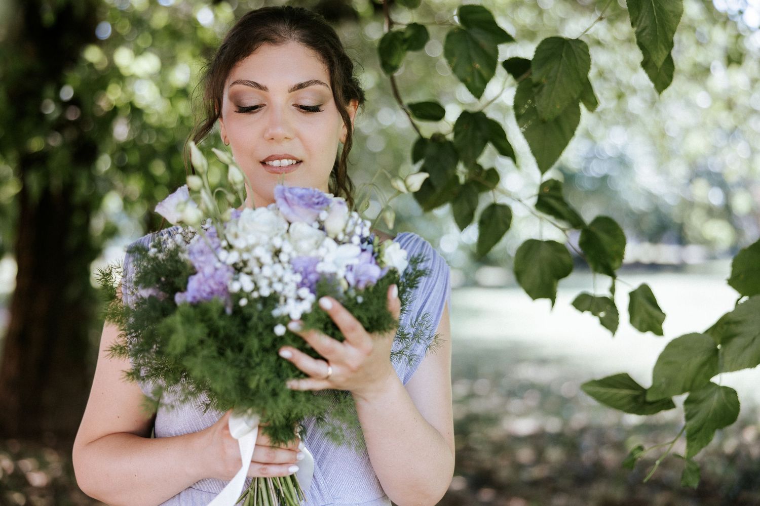 A bride in a white gown poses outdoors with a natural green bouquet in a garden setting.