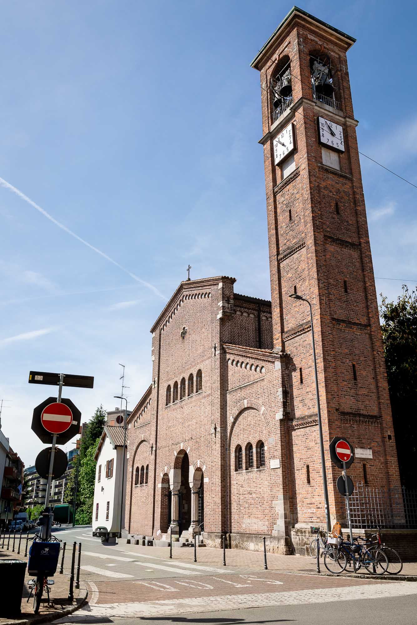 Brick Romanesque church with tall clock tower and arched entrance under blue sky, street signs visible in foreground.