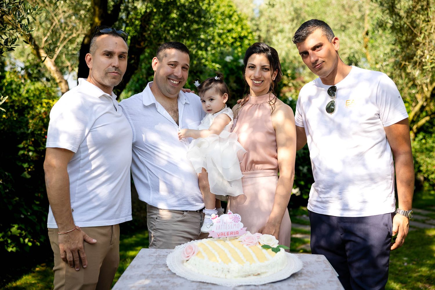 Family group of five adults and baby girl celebrating first birthday with white cake in garden setting.