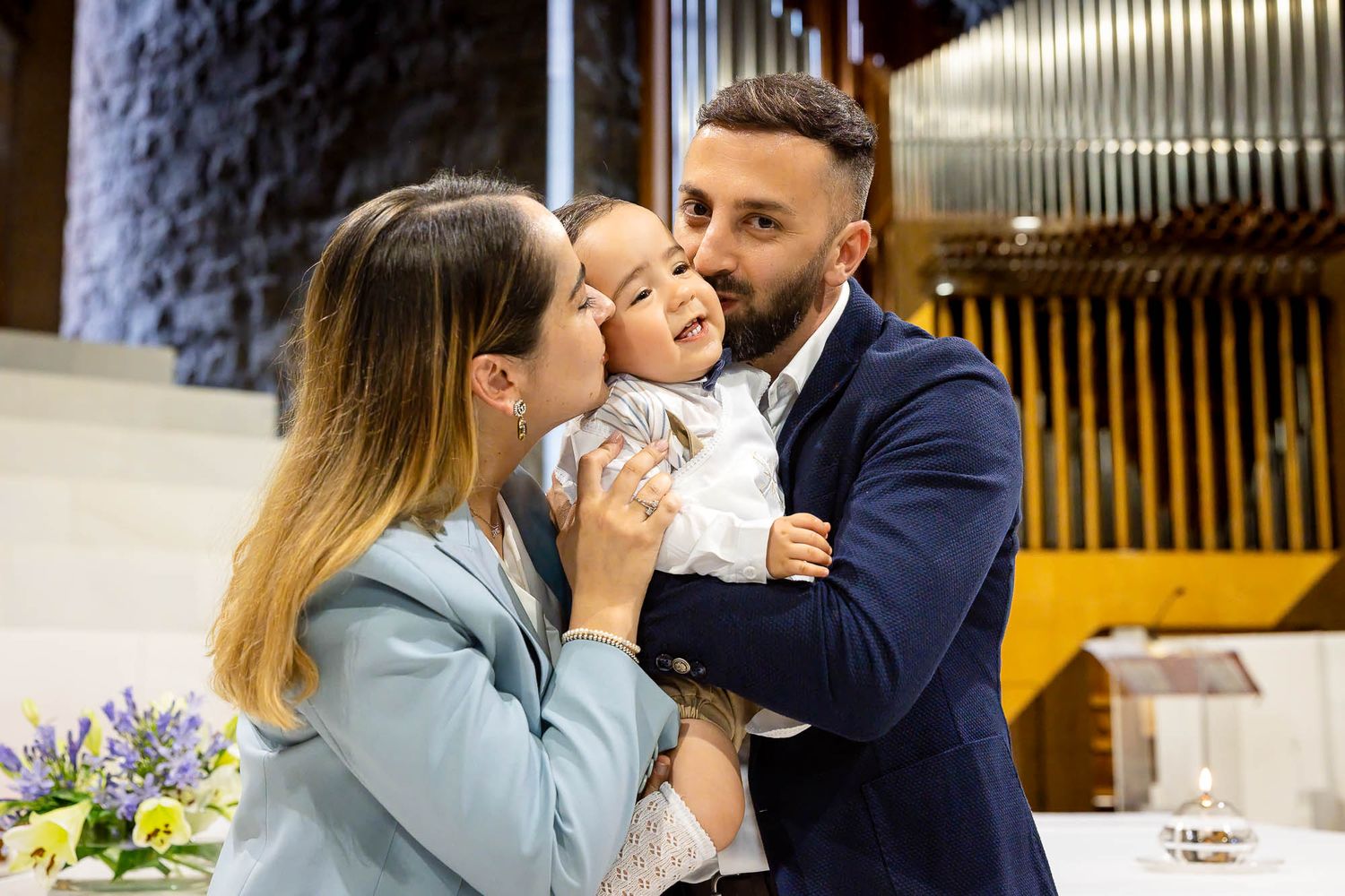 Happy family with parents holding smiling baby in church during baptism ceremony.