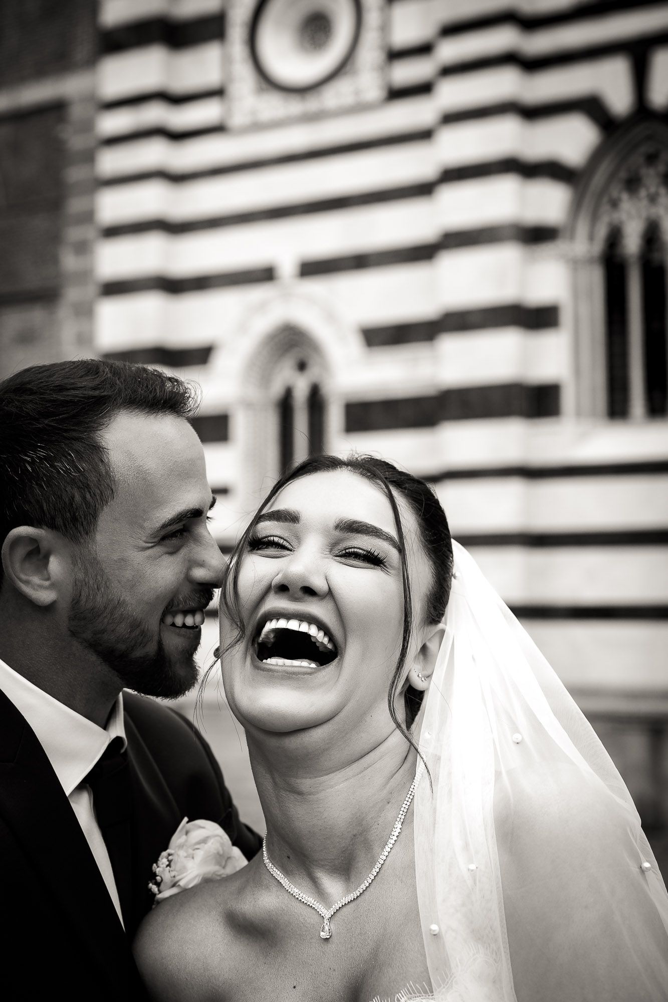 Black and white photo of laughing bride and groom outside ornate church on wedding day.