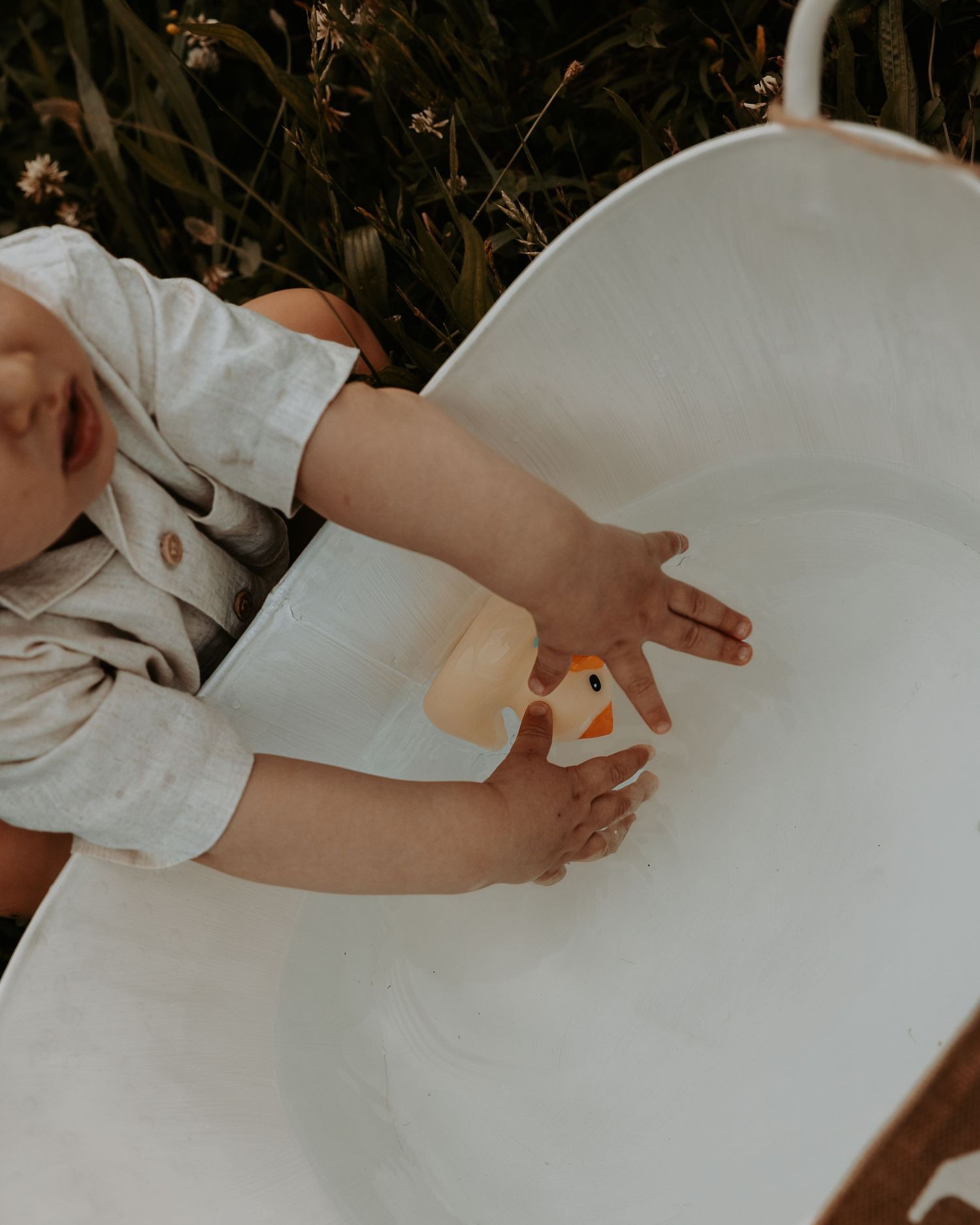 Baby lying in a white Moses basket outdoors, reaching for a small toy duck surrounded by lush green foliage.