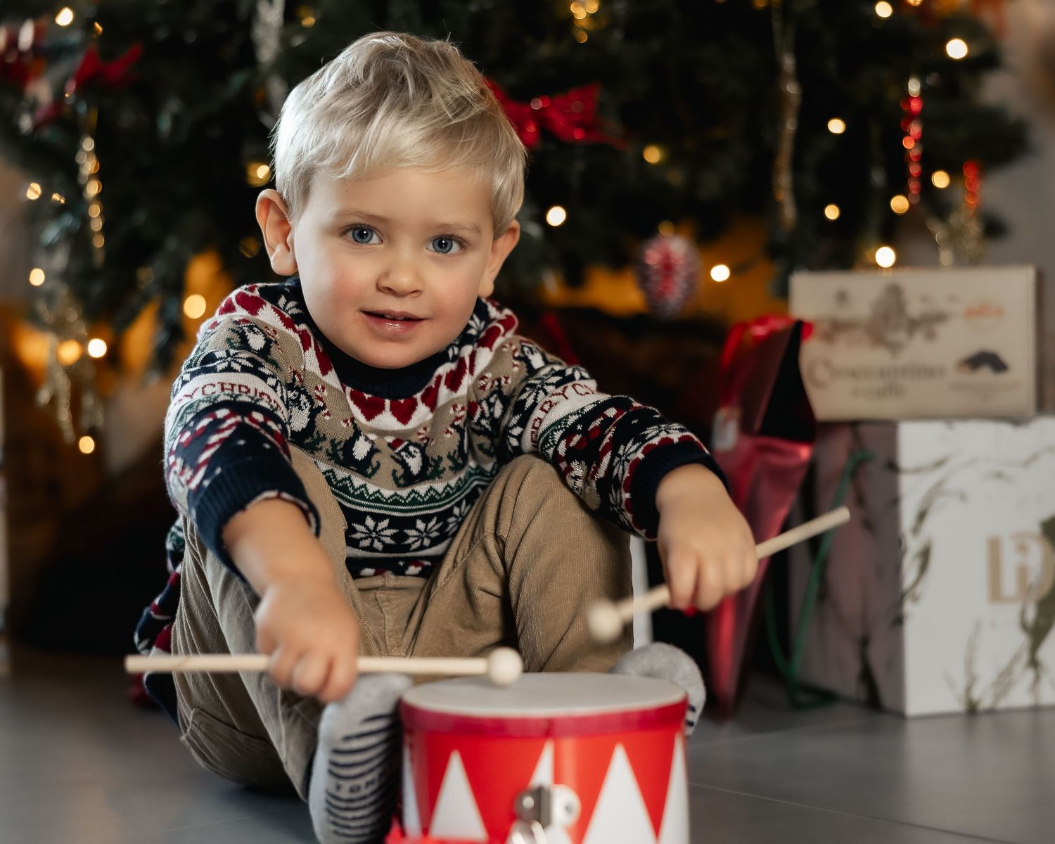 Young boy in festive sweater playing toy drum near Christmas tree with bokeh lights and gifts.