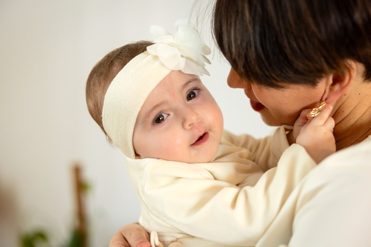 A mother in cream clothing holds her adorable baby girl wearing a white floral headband, sharing a tender moment.