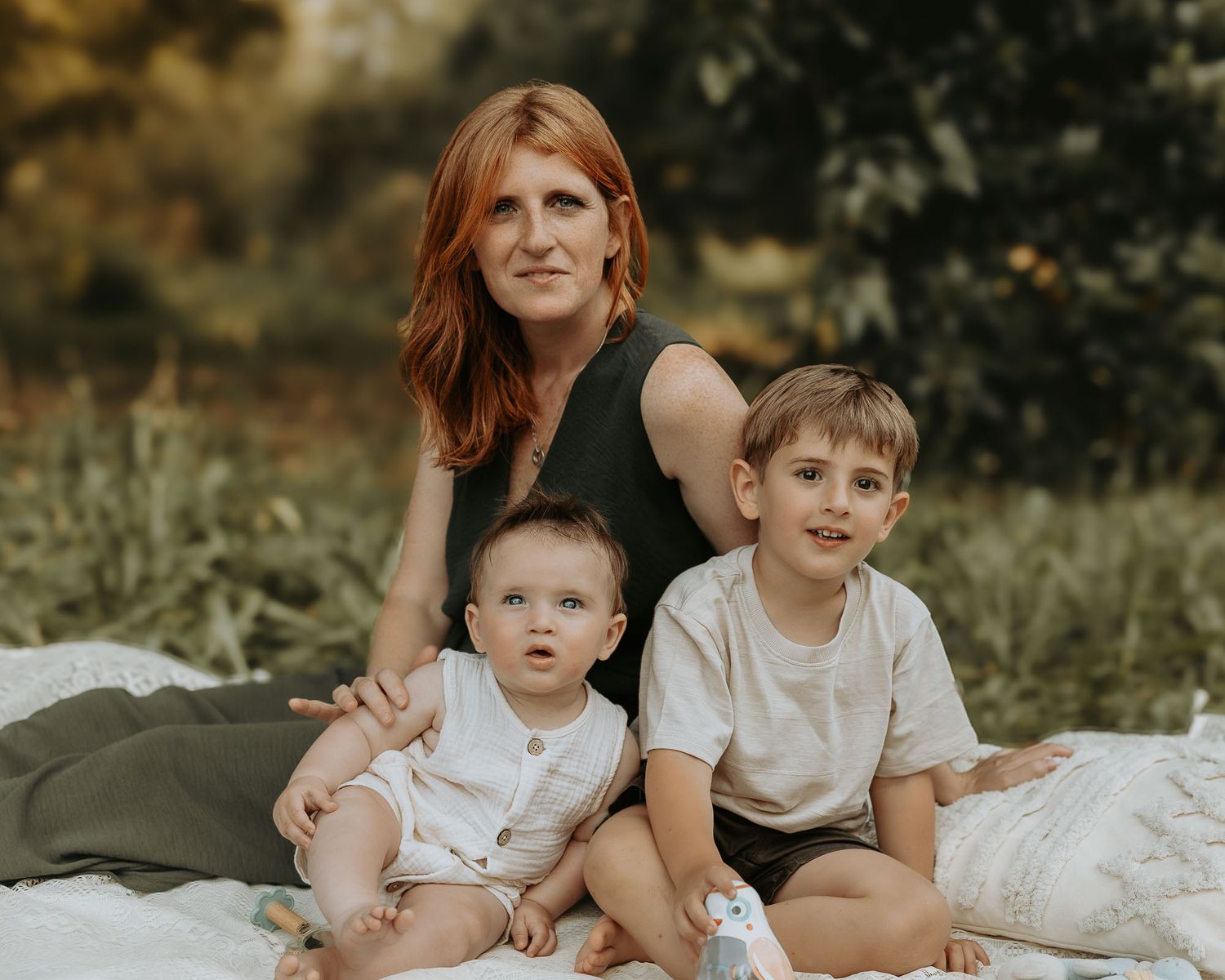 A red-haired woman sits outdoors on a blanket with two young boys in a lush green park setting.