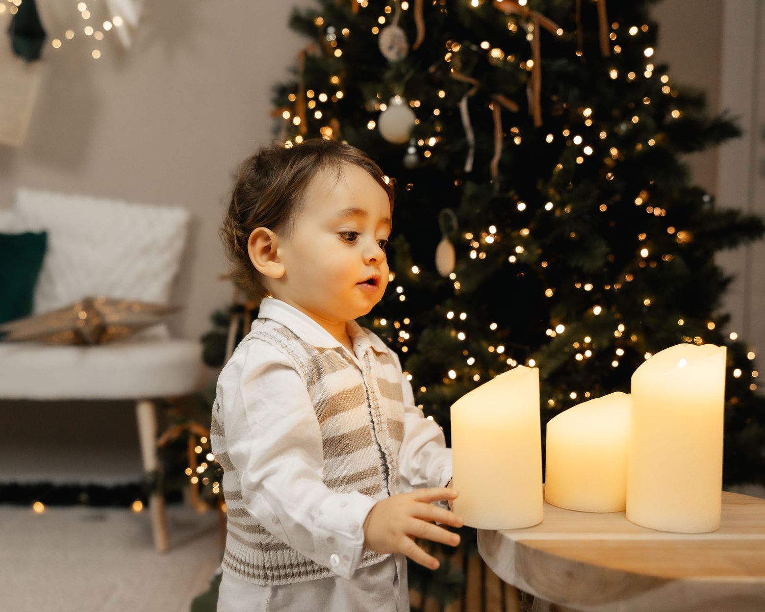 Toddler in festive outfit admiring glowing white candles near a decorated Christmas tree with warm lights.