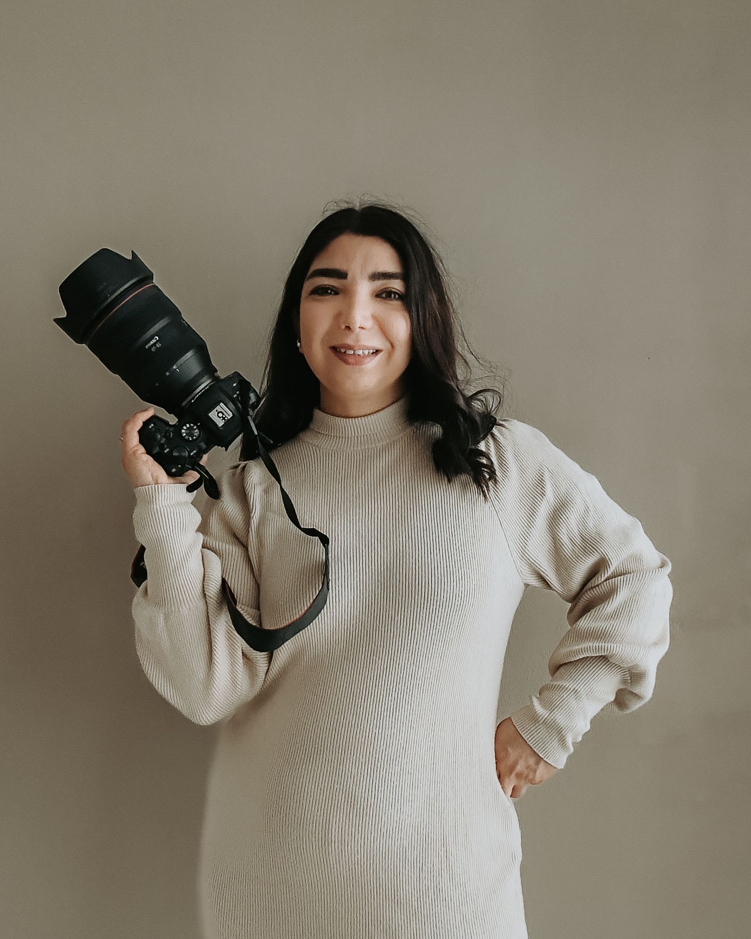 Professional photographer in a cream sweater holding a DSLR camera against neutral background.