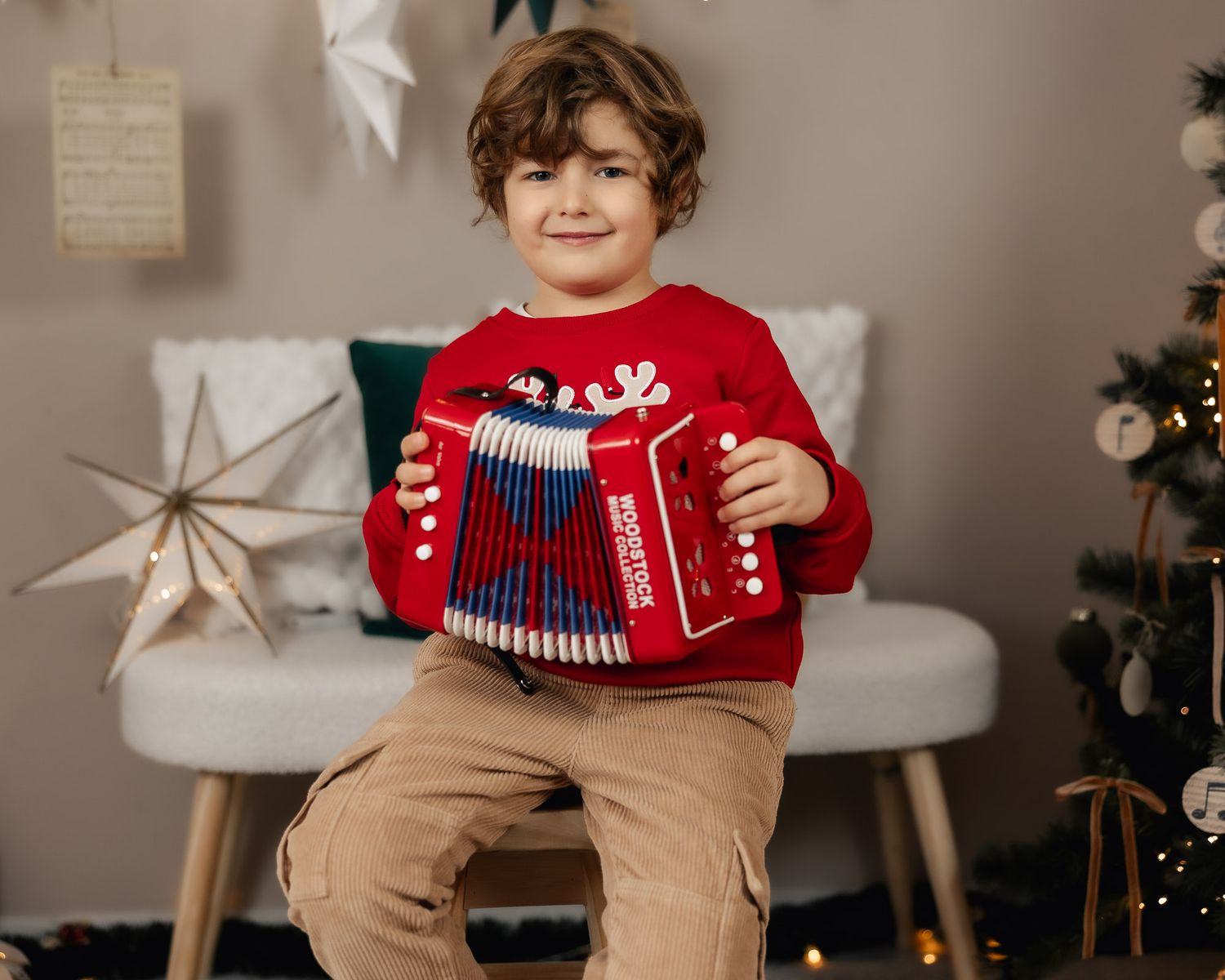 Young boy in red reindeer sweater plays accordion in festive Christmas decorated room with tree.
