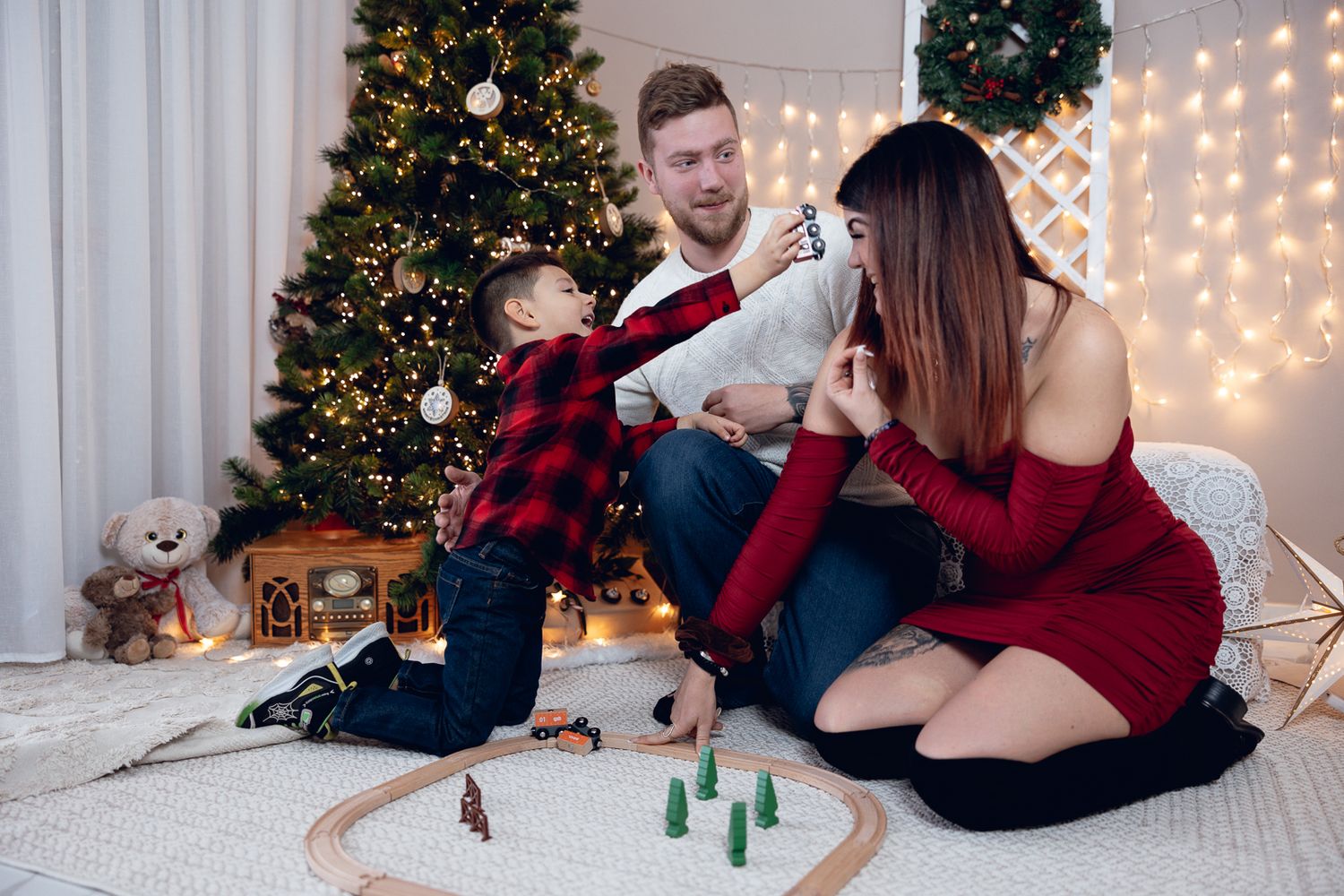 Family in matching red and black outfits poses for holiday photos in front of Christmas tree and twinkle lights.