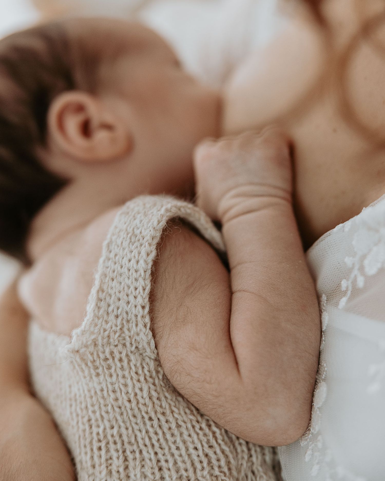 Close-up of a newborn baby in a knit romper breastfeeding, with tiny fist visible against mother's chest.