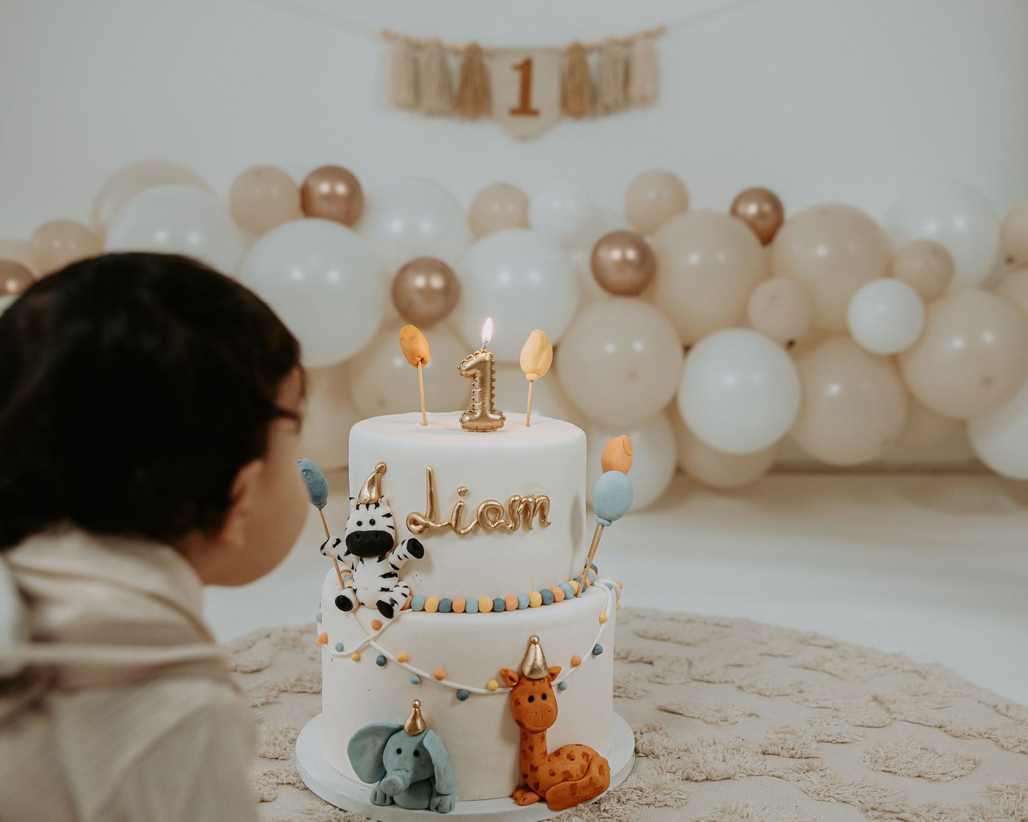 Child looks at a first birthday cake named Liam with animal figurines, surrounded by beige and gold balloon decorations.