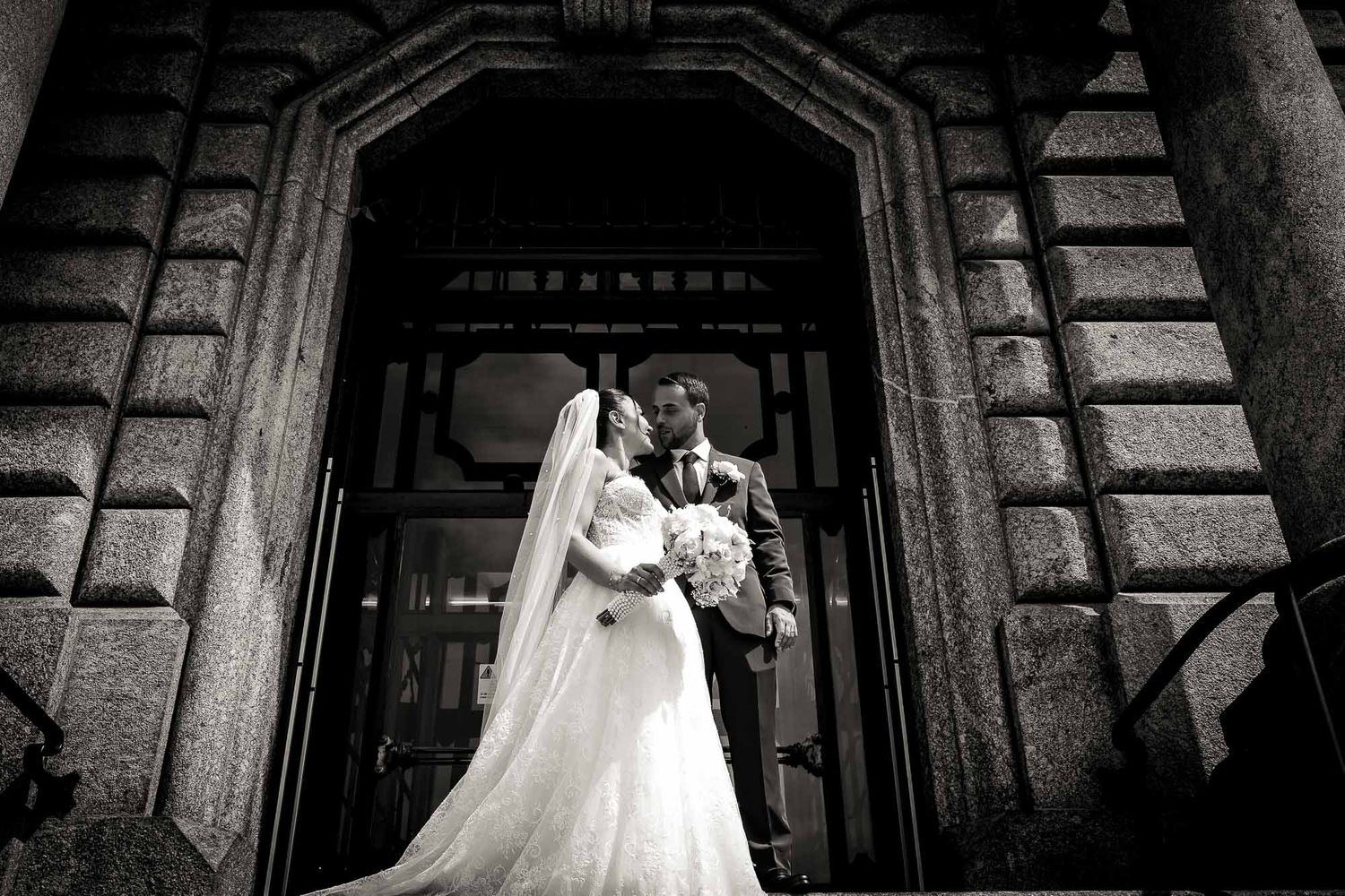 Black and white wedding photo of bride and groom kissing at ornate building entrance, bride in elegant gown with veil.