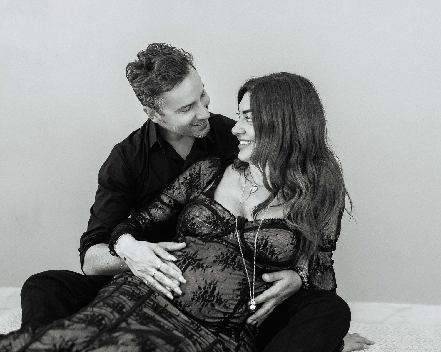 Black and white maternity photo of smiling couple sitting together, man embracing pregnant woman from behind.