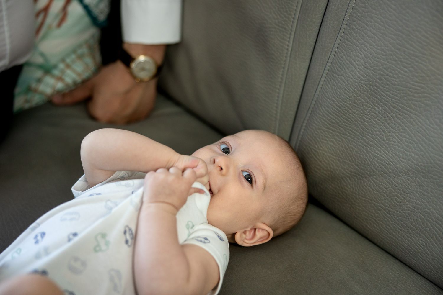 A cute baby with blue eyes lying on a gray sofa, sucking fingers while an adult sits nearby.