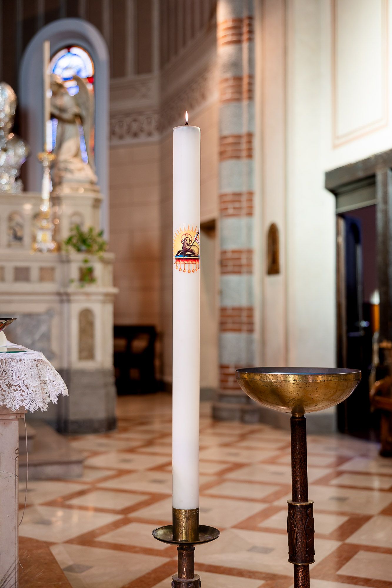 Tall white paschal candle with religious emblem on brass stand inside ornate Catholic church with marble altar.