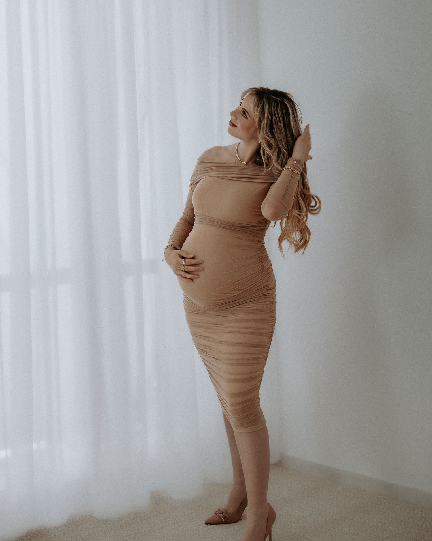 Una donna incinta, con un elegante abito midi color beige/tan arricciato e con spalle scoperte durante servizio fotografico in studio a mila