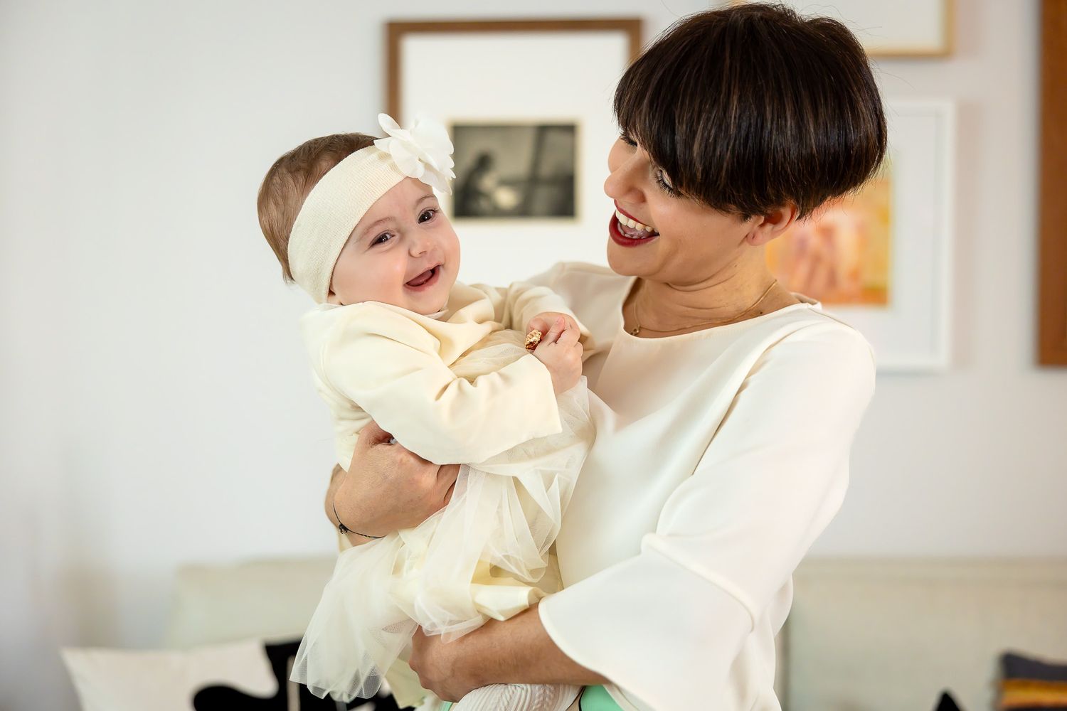 A smiling mother in a white top holds her happy baby girl wearing a white dress and headband indoors.