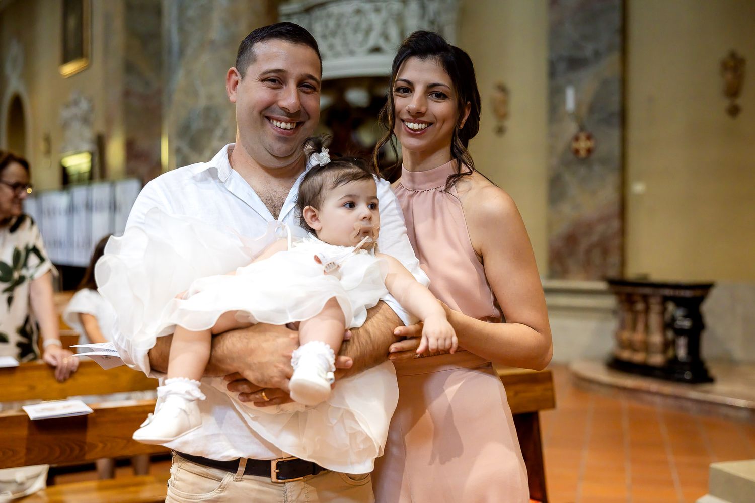A couple smiles holding their baby in a white christening gown inside a church during a baptism ceremony.