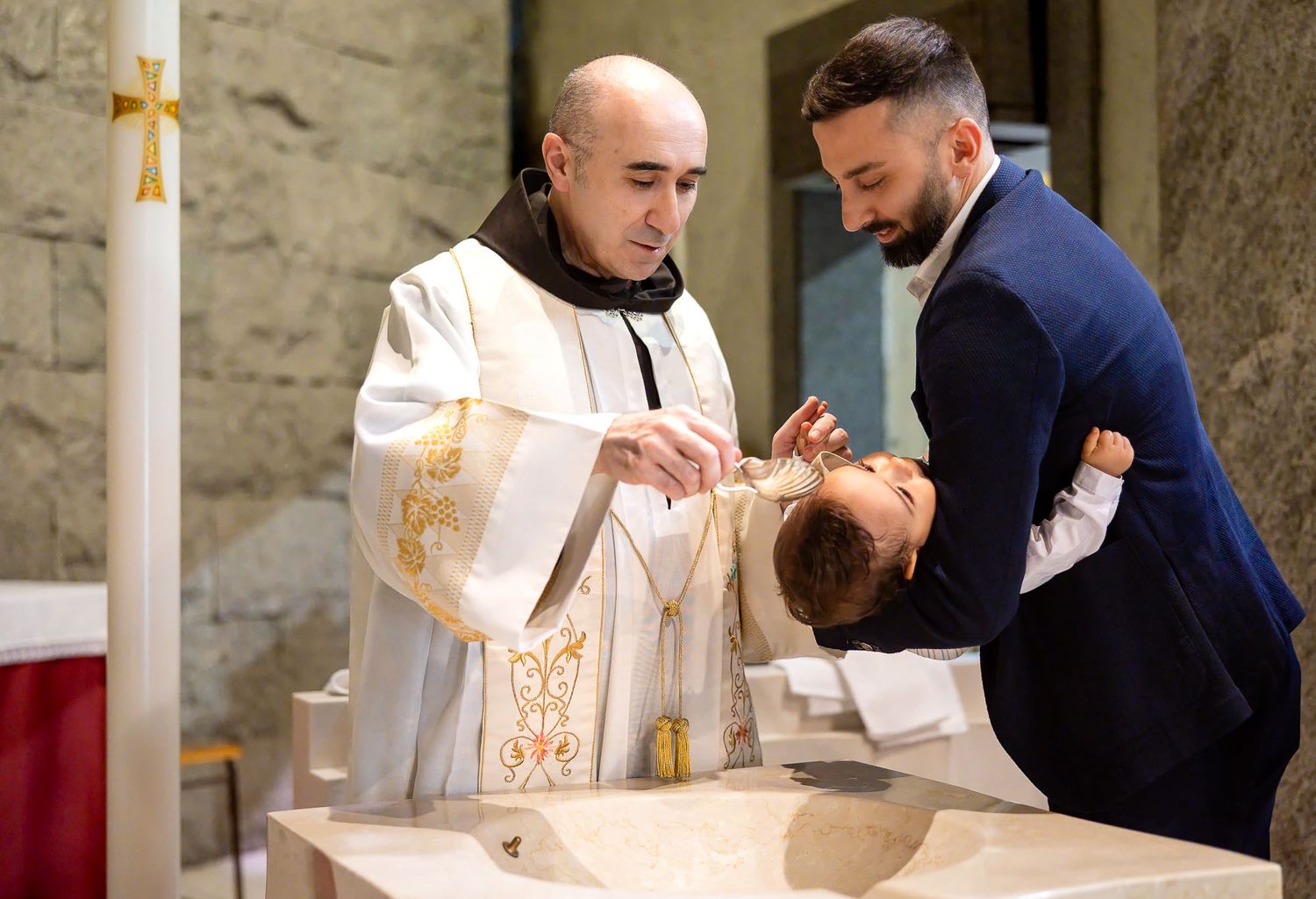 Priest performing a baptism ceremony, pouring holy water over an infant held by a man in a blue suit.