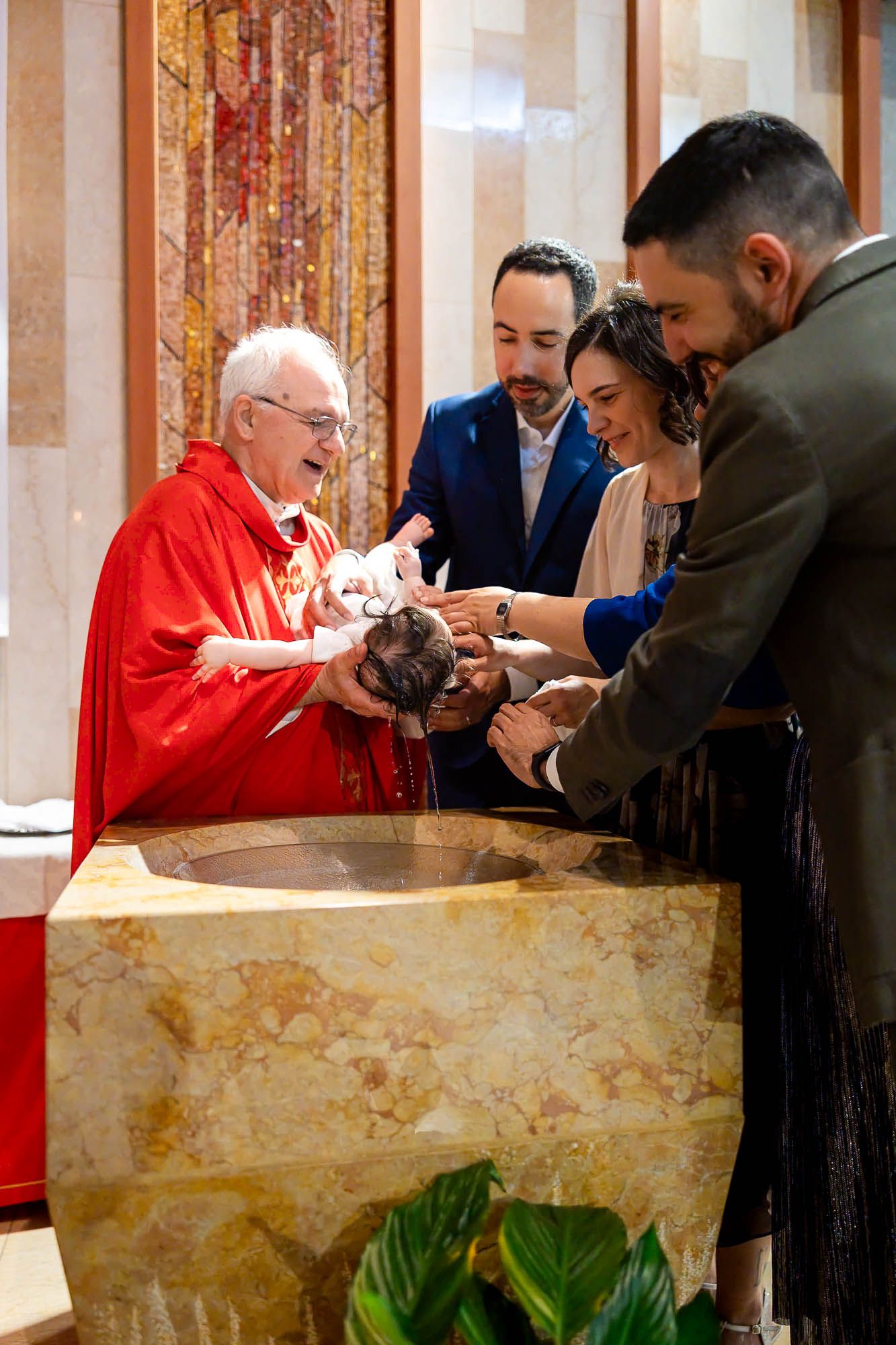 Priest in red vestments performs infant baptism at font with parents and godfather in church.