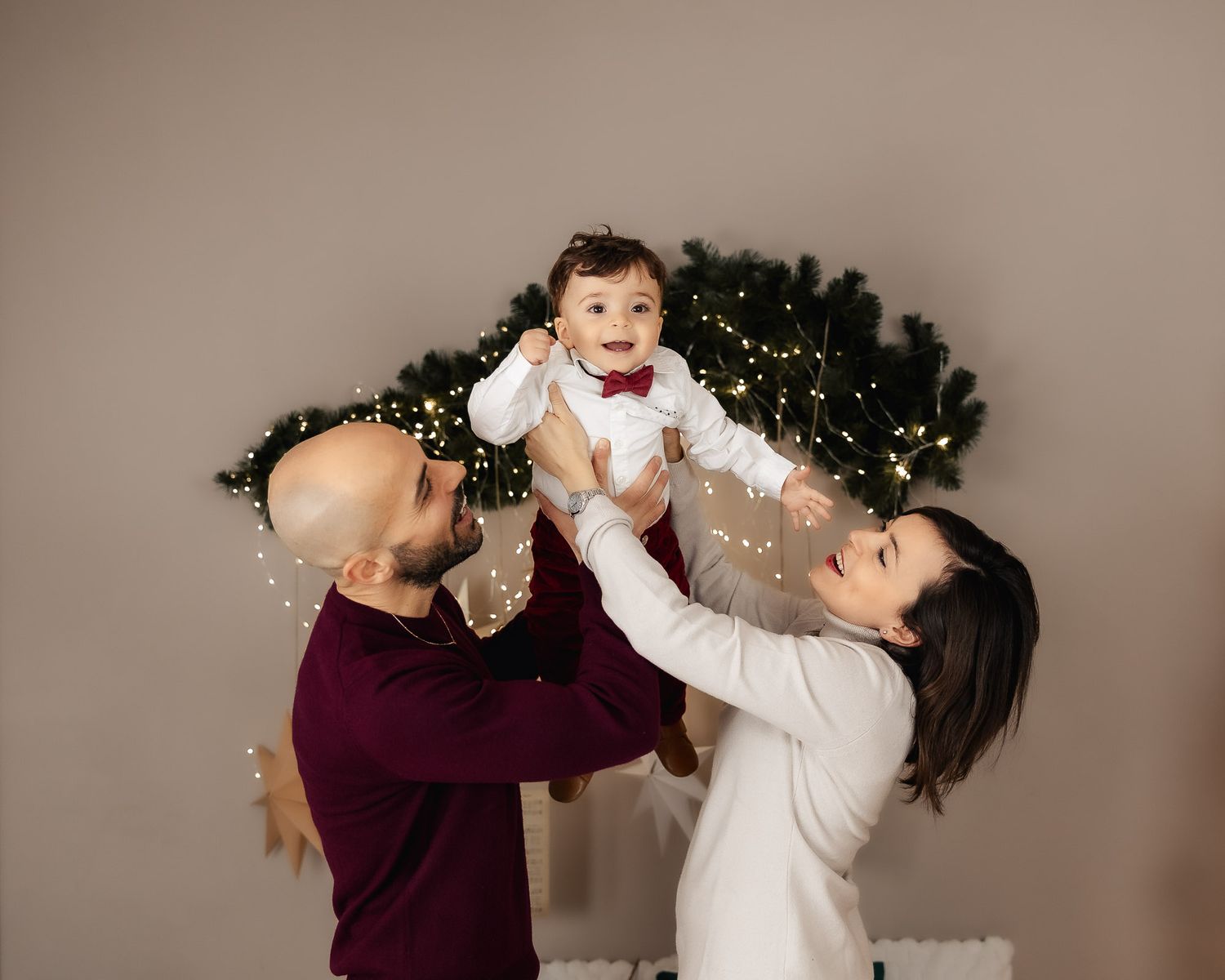 Parents joyfully lifting smiling baby in bow tie near Christmas tree with fairy lights in festive holiday photo.