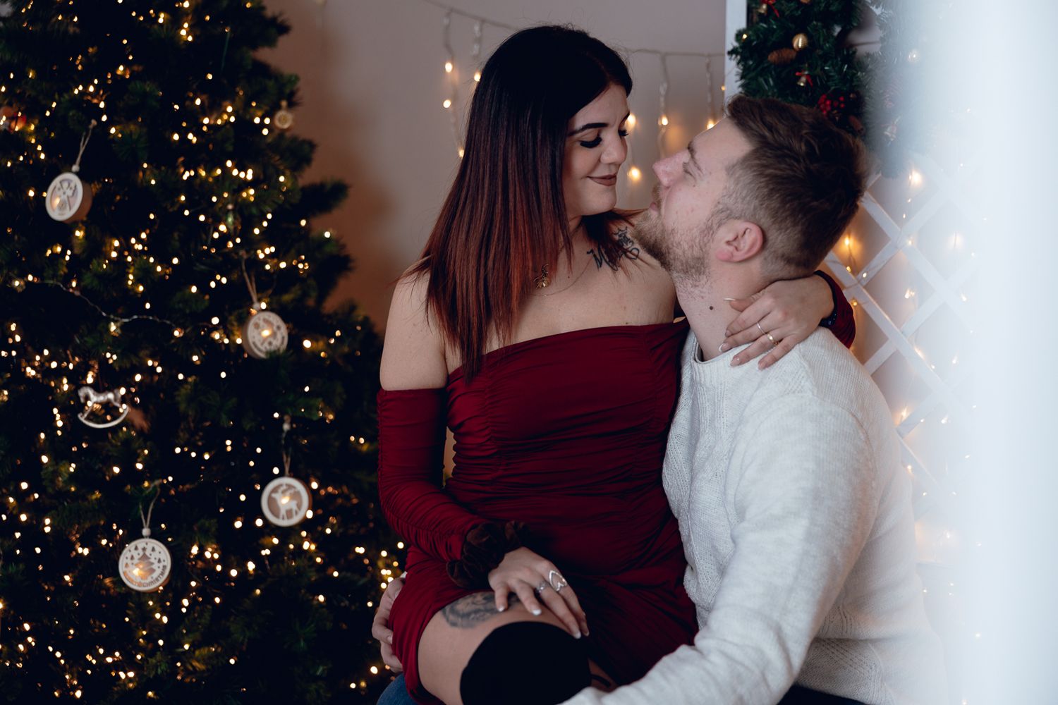 A romantic couple embraces near a decorated Christmas tree with twinkling lights in the background.