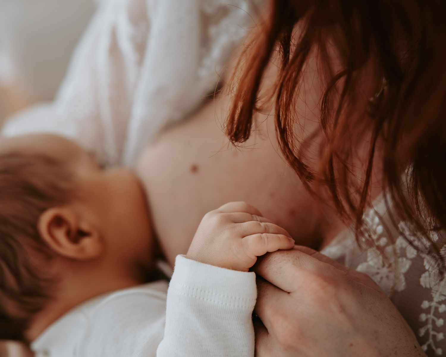 A mother with red hair breastfeeding her newborn baby dressed in white, captured in soft natural light.