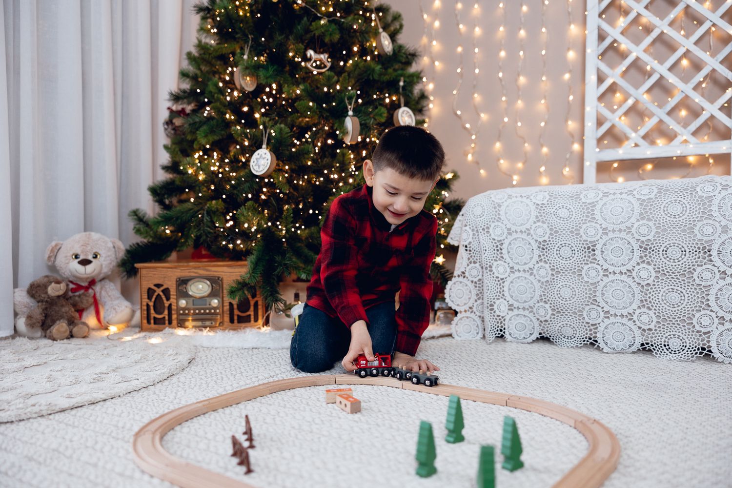 Child playing with wooden toy train set near Christmas tree with twinkling lights in background.