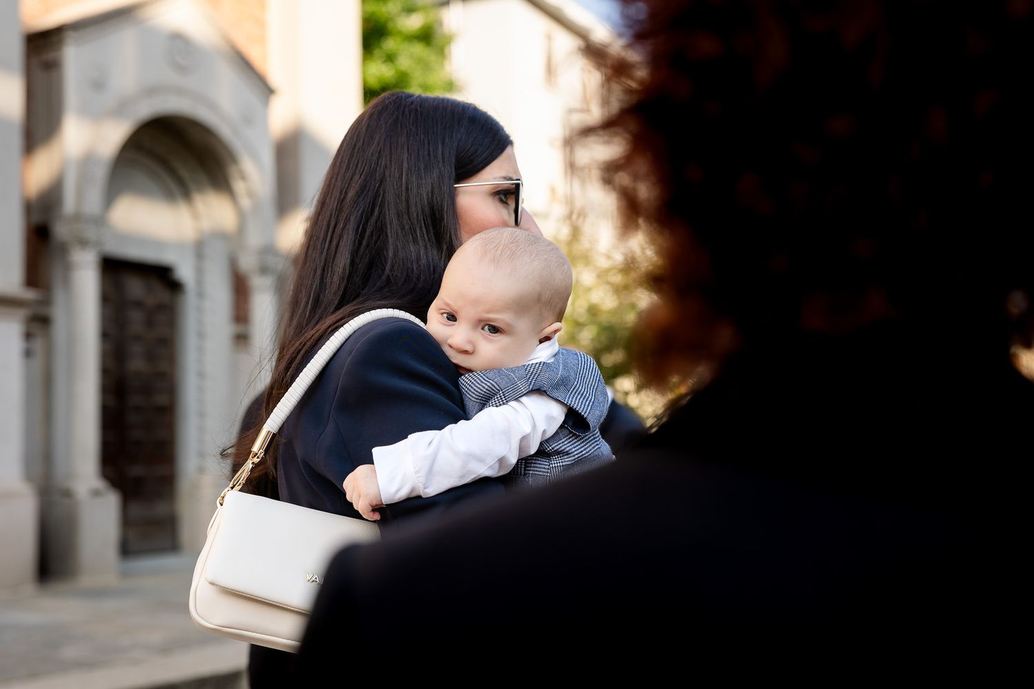 A mother tenderly holds her baby outside a historic building, wearing a white handbag in soft natural light.