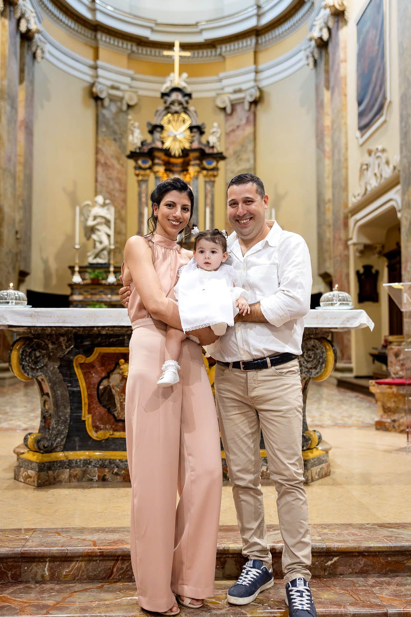 Family posing in ornate church during baptism ceremony, mother in pink jumpsuit holding baby in white outfit.