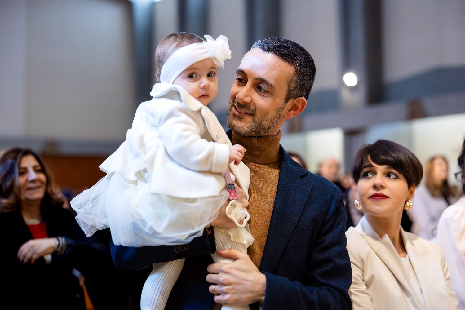 A man holds a baby dressed in white at a christening ceremony, with a woman smiling nearby in a church.