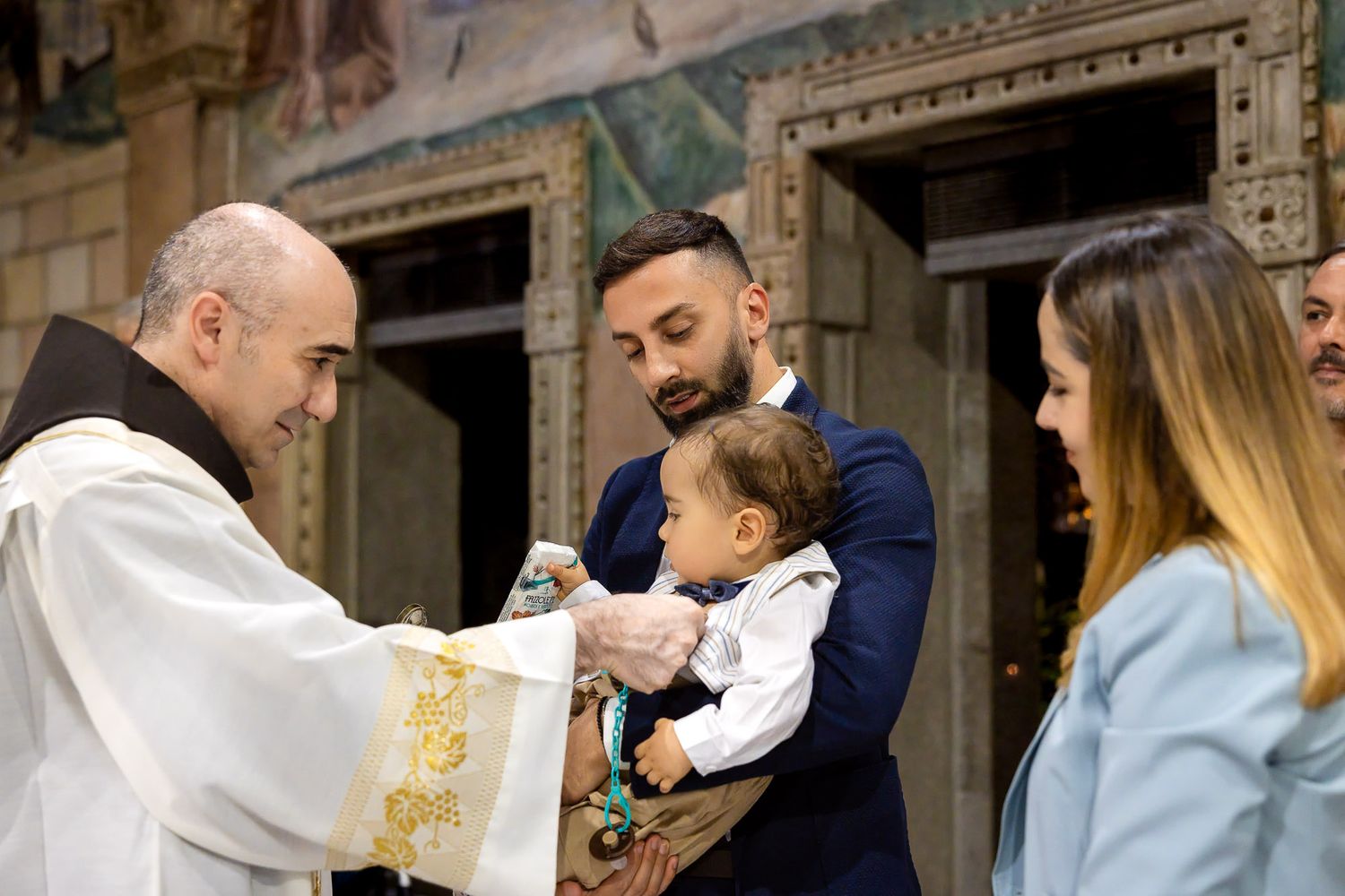 Catholic priest performing a baptism ritual with parents holding their infant baby in a decorated church.