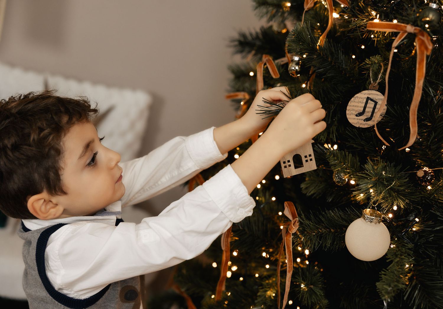 Young boy decorating a Christmas tree with ornaments and golden ribbons, wearing a white shirt during holiday season.