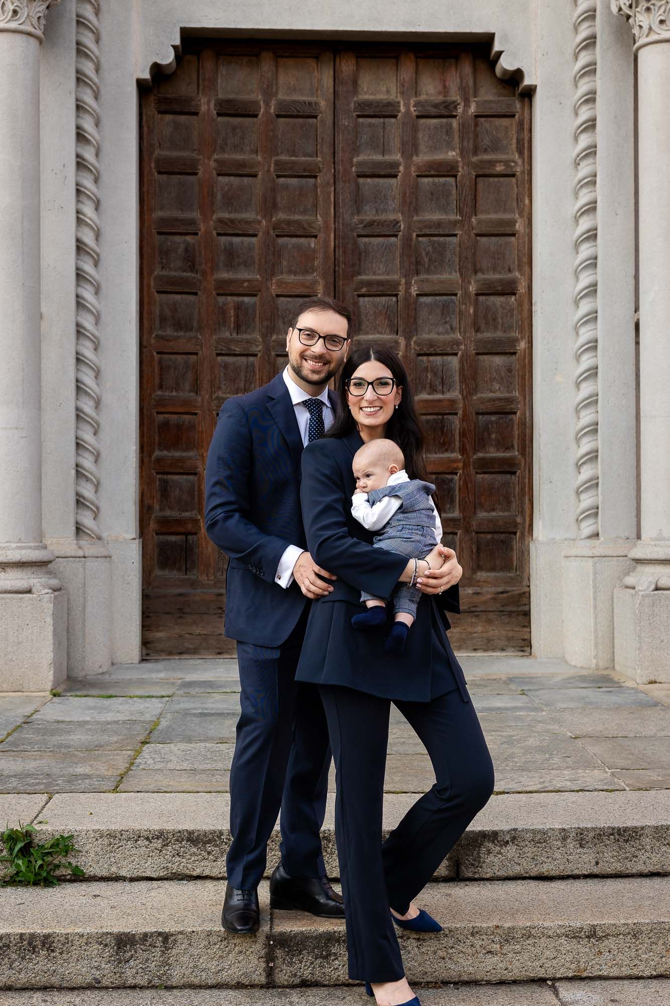 A smiling couple in navy suits holding a baby on stone steps before a large wooden door.