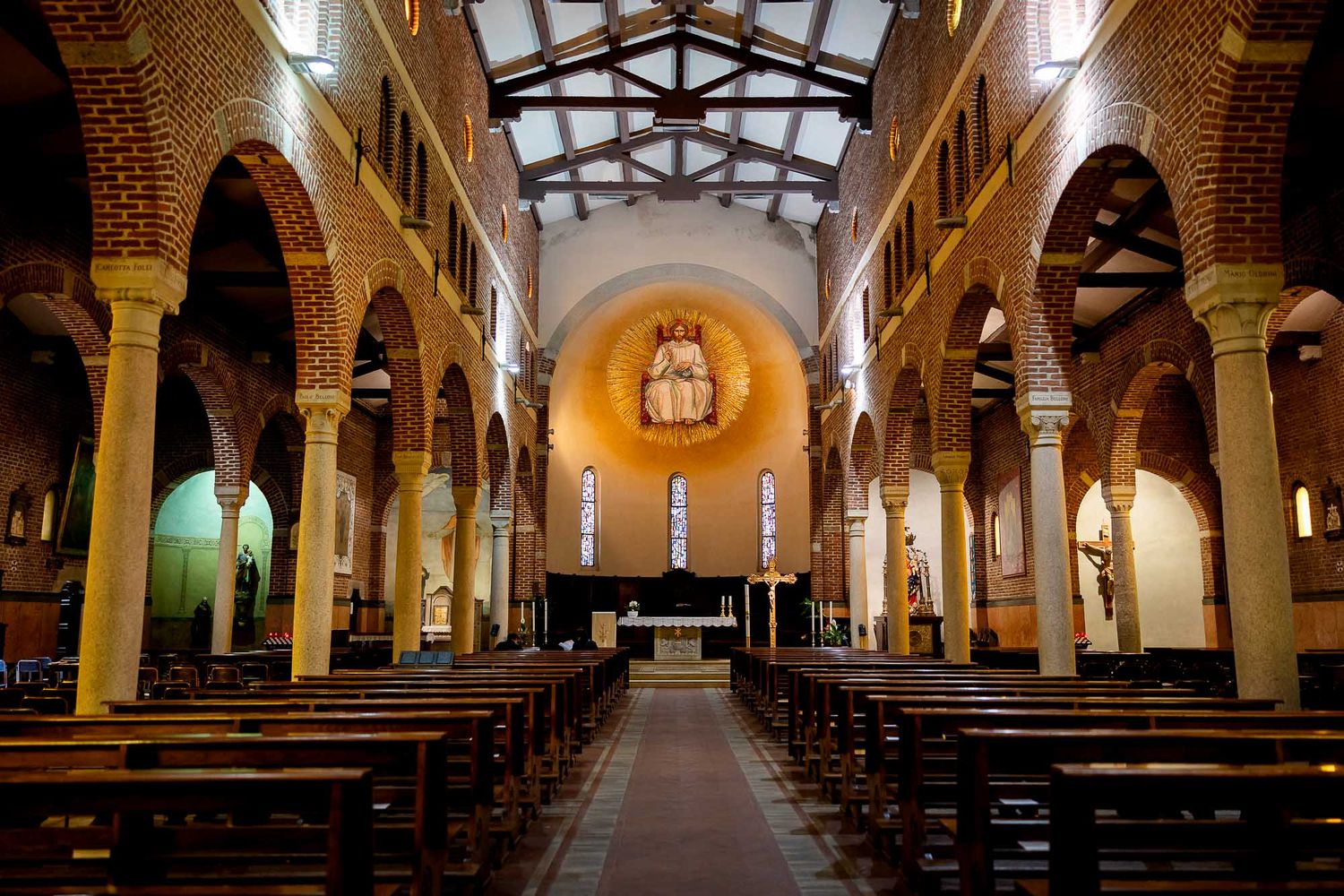 Interior of a historic brick church with arched columns, wooden pews, and a glowing altar mosaic.