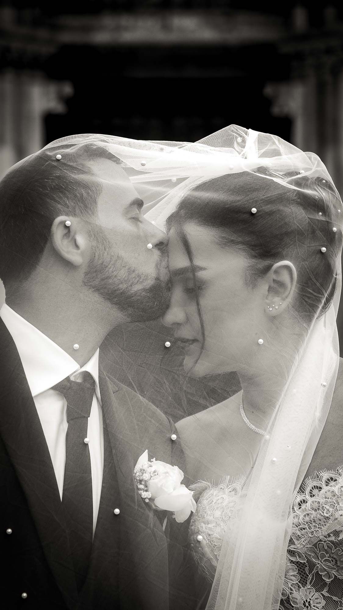 Black and white wedding photo of groom kissing bride on cheek under pearl-adorned veil.