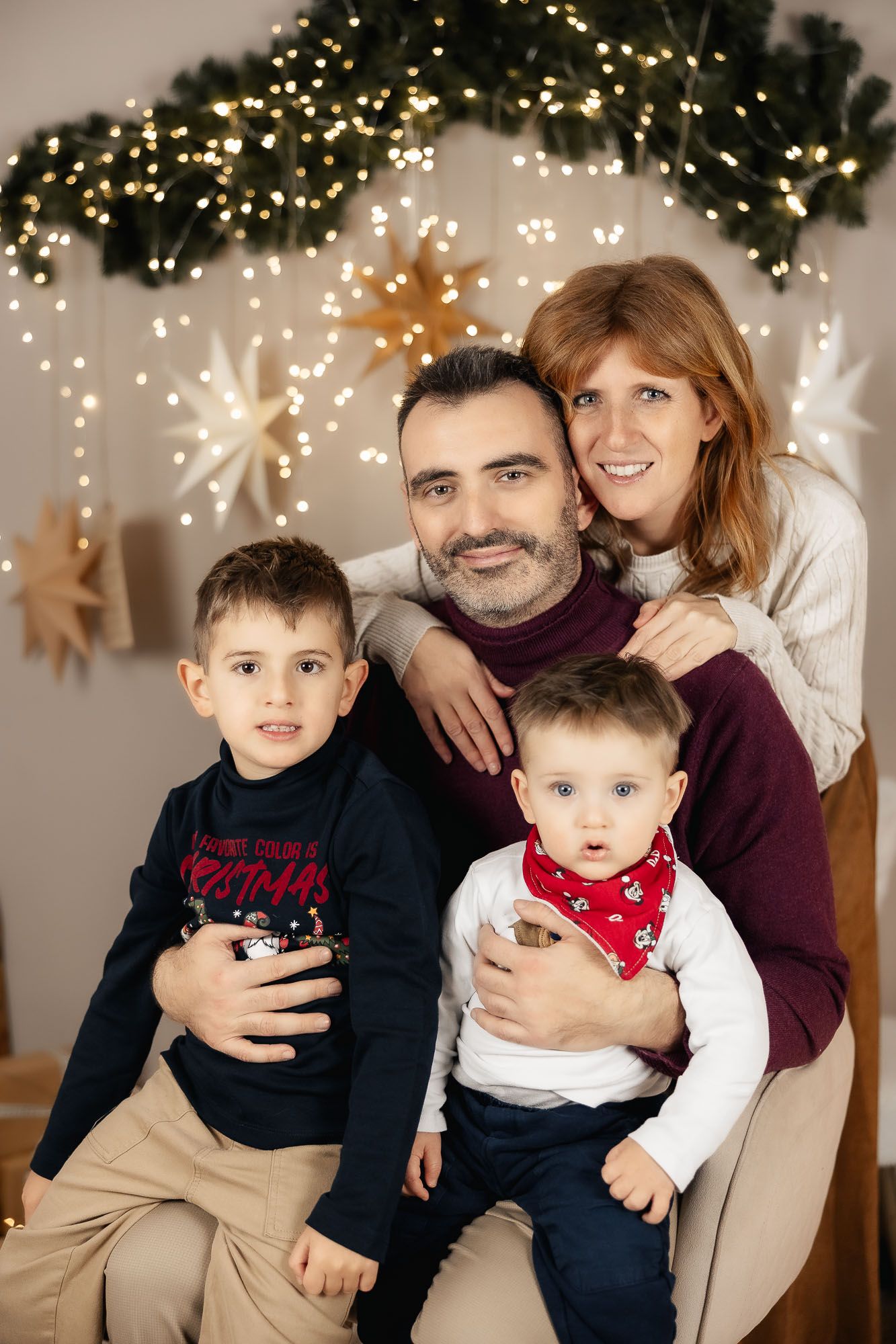 Family of four posing for a festive Christmas portrait with holiday lights and star decorations in the background.