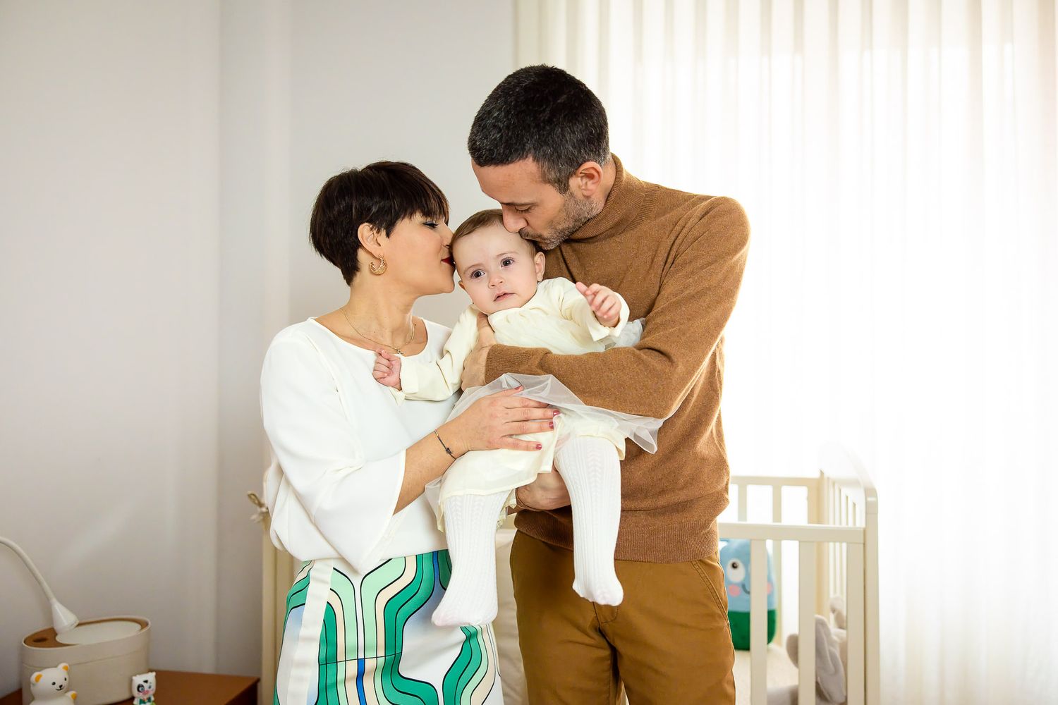 A couple lovingly holds their baby in a bright nursery, with the man kissing the infant's head.