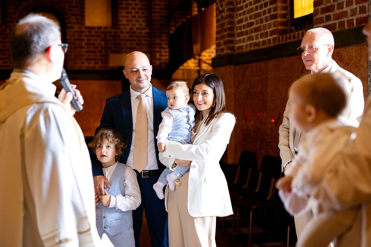 Family gathered in a church for a baptism ceremony, with a priest, parents holding a baby, and young children present.