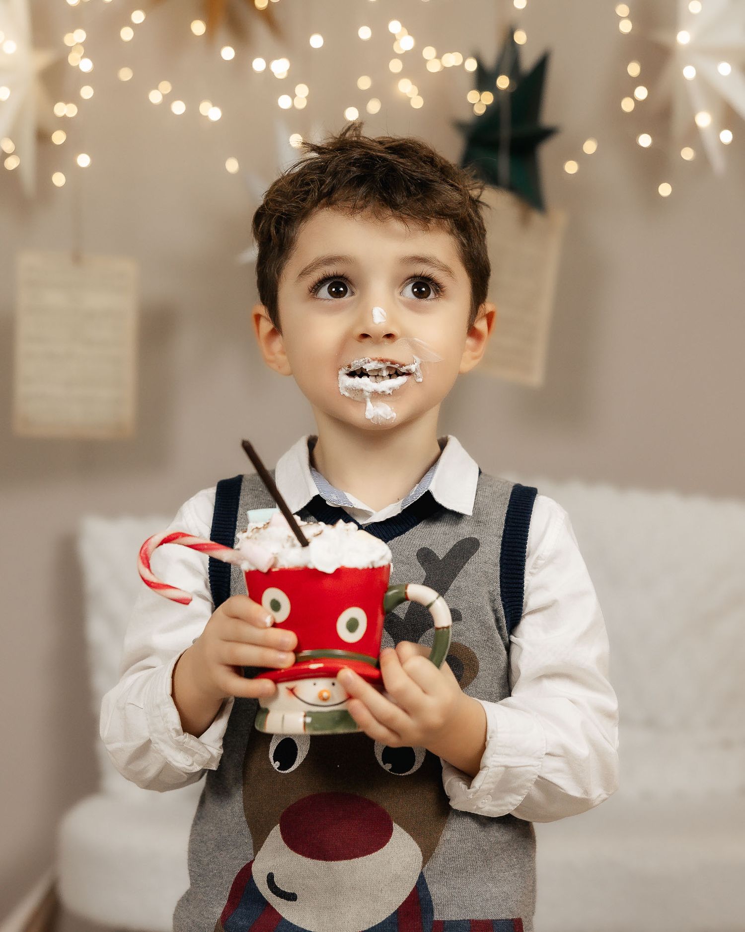 A young boy in a reindeer sweater holds a festive red mug with whipped cream on his face near Christmas lights.