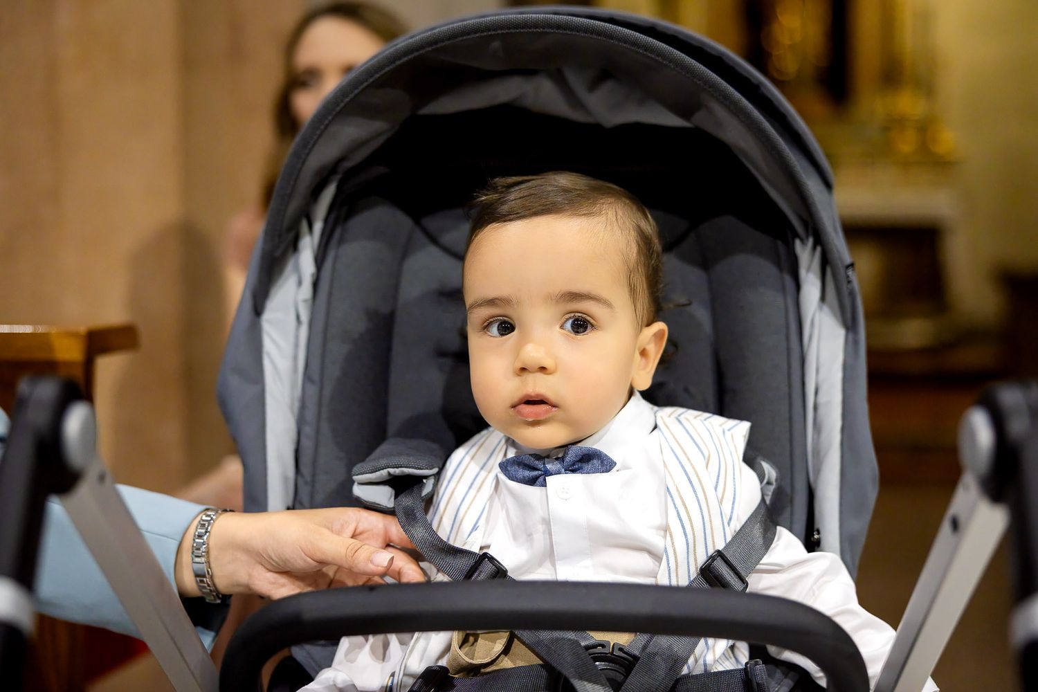 Adorable baby boy in a blue bow tie sitting in a gray stroller during a christening ceremony.