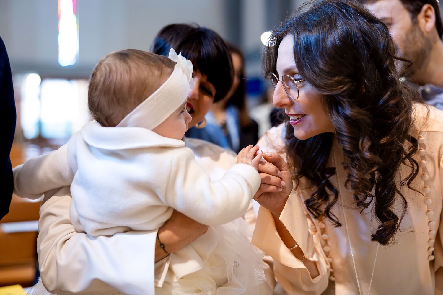 A smiling woman with curly hair holds hands with a baby in a white christening gown during a baptism ceremony in a church.