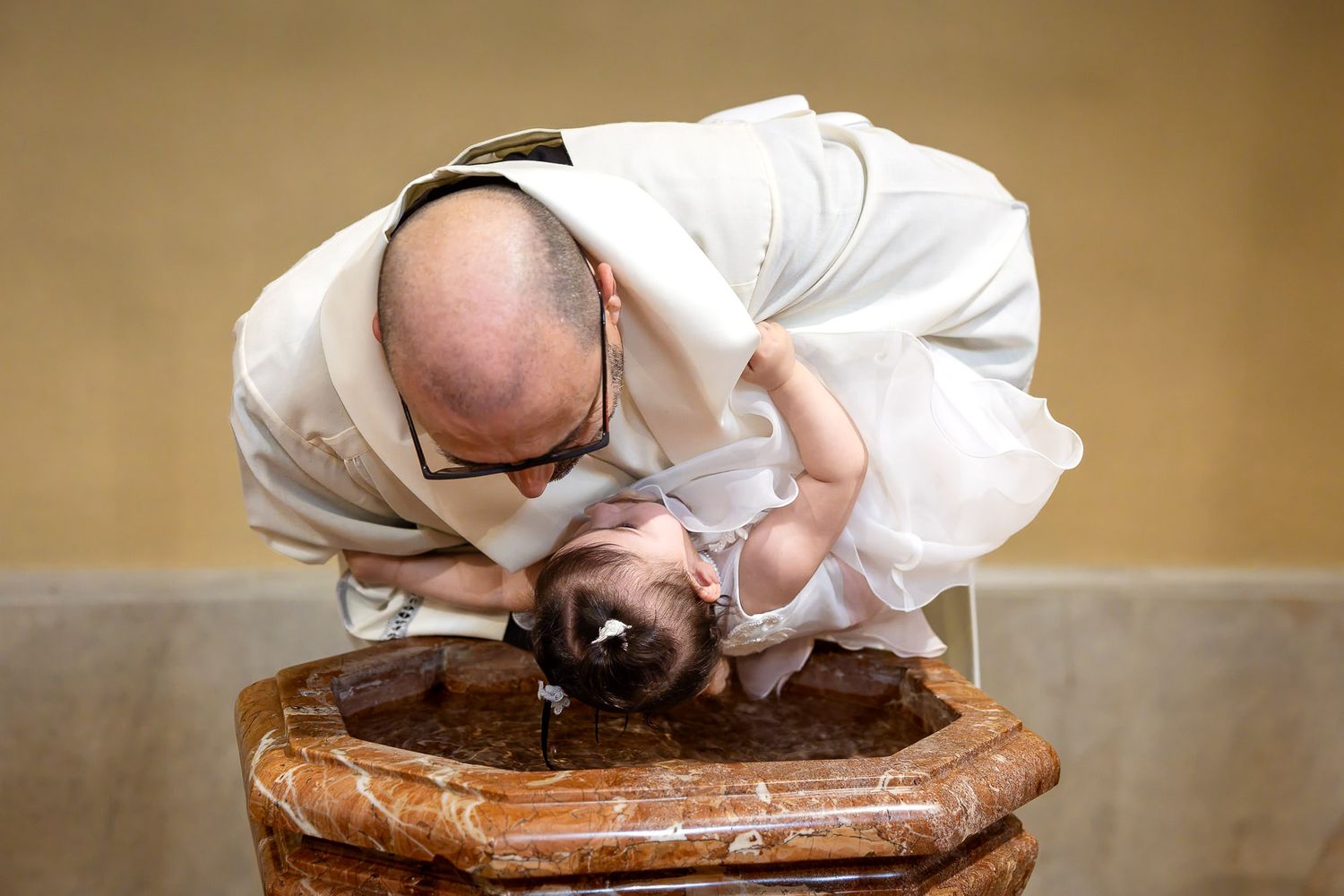 Priest baptizing a baby at a stone font during a Catholic baptism ceremony.