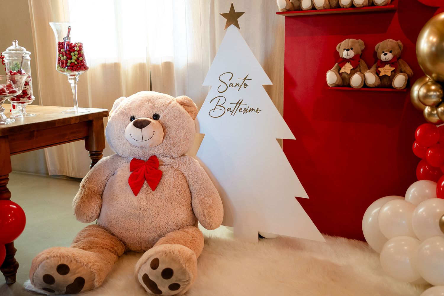 Large teddy bear with red bow sits beside a white Christmas tree sign reading Santa Balloons, with festive red and white decor.
