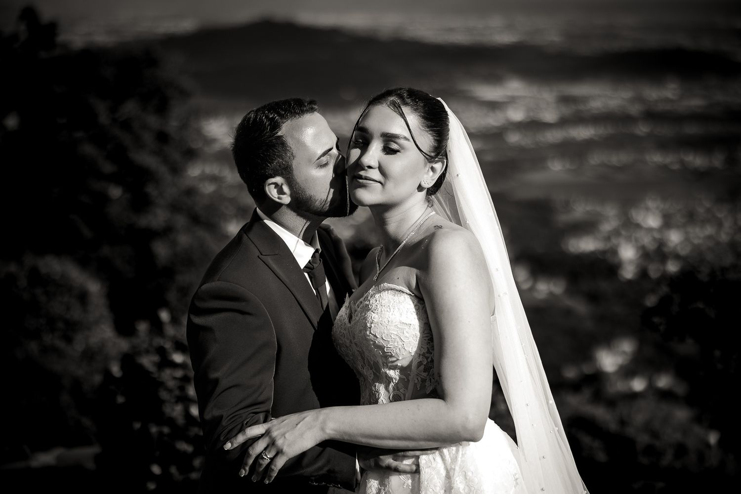 Black and white wedding photo of groom kissing bride on cheek with city lights in background.