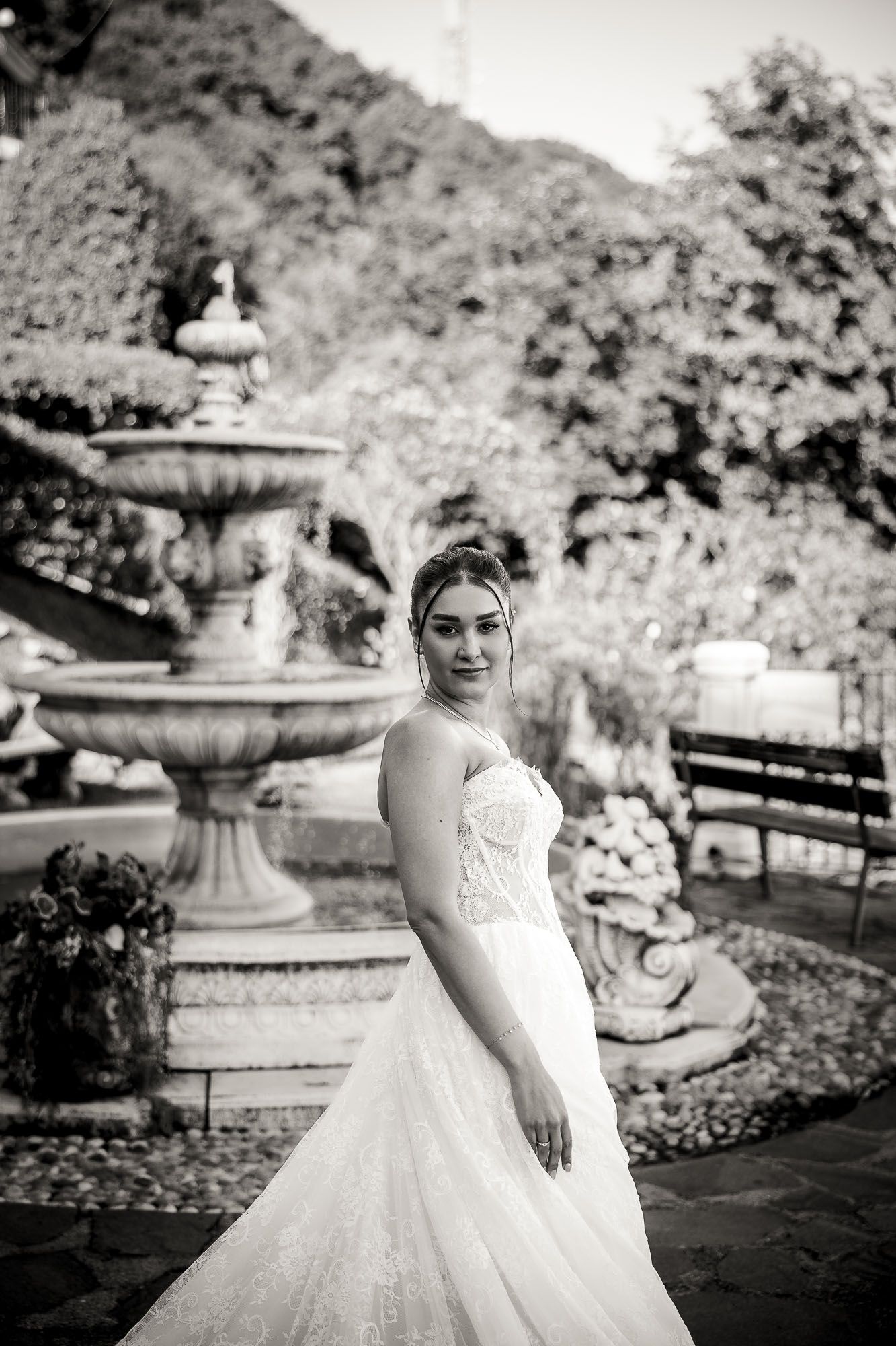 Bride in lace wedding gown poses before stone fountain in garden, black and white bridal portrait.