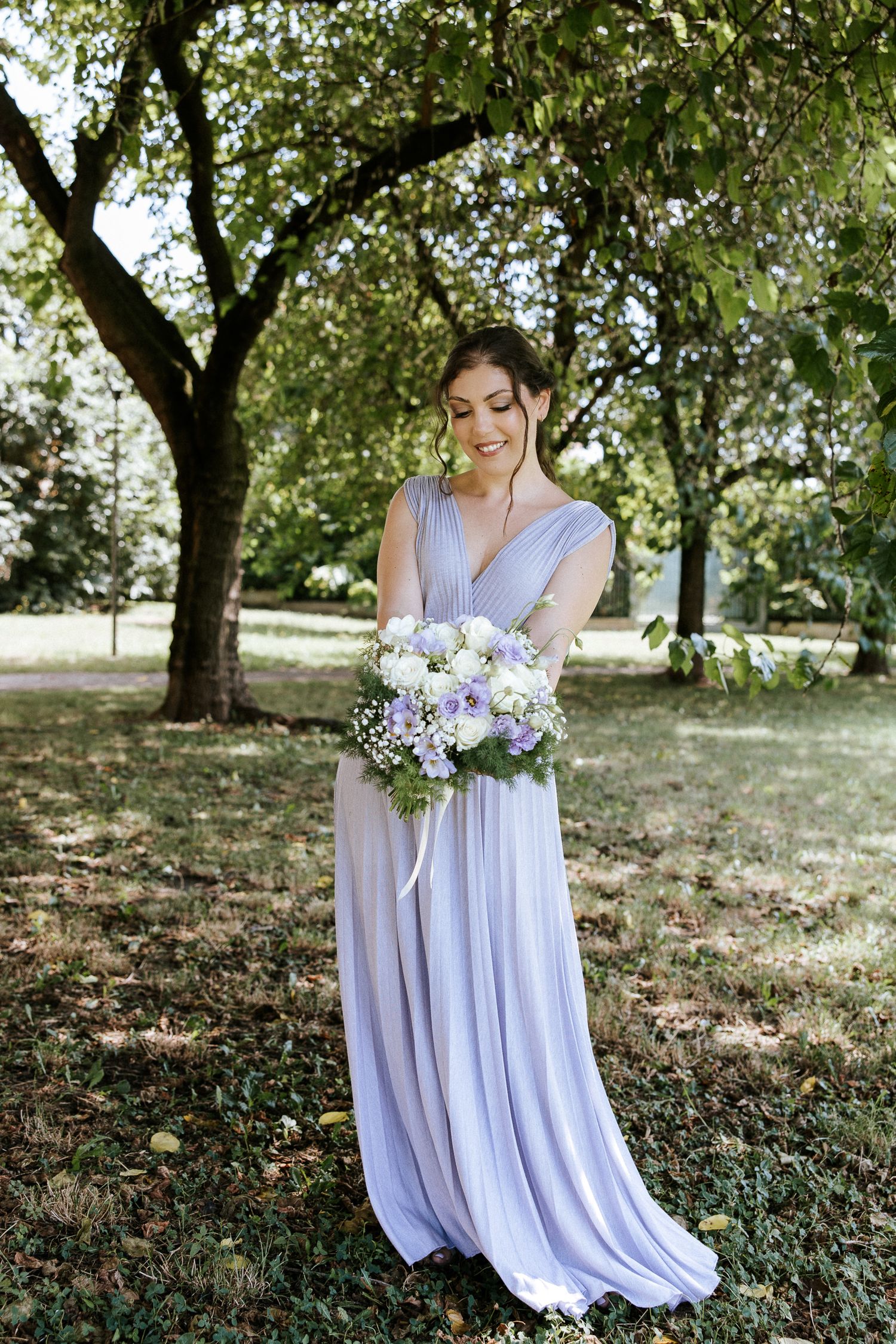 A flowing lavender dress is shown against a natural garden backdrop with trees and dappled sunlight.