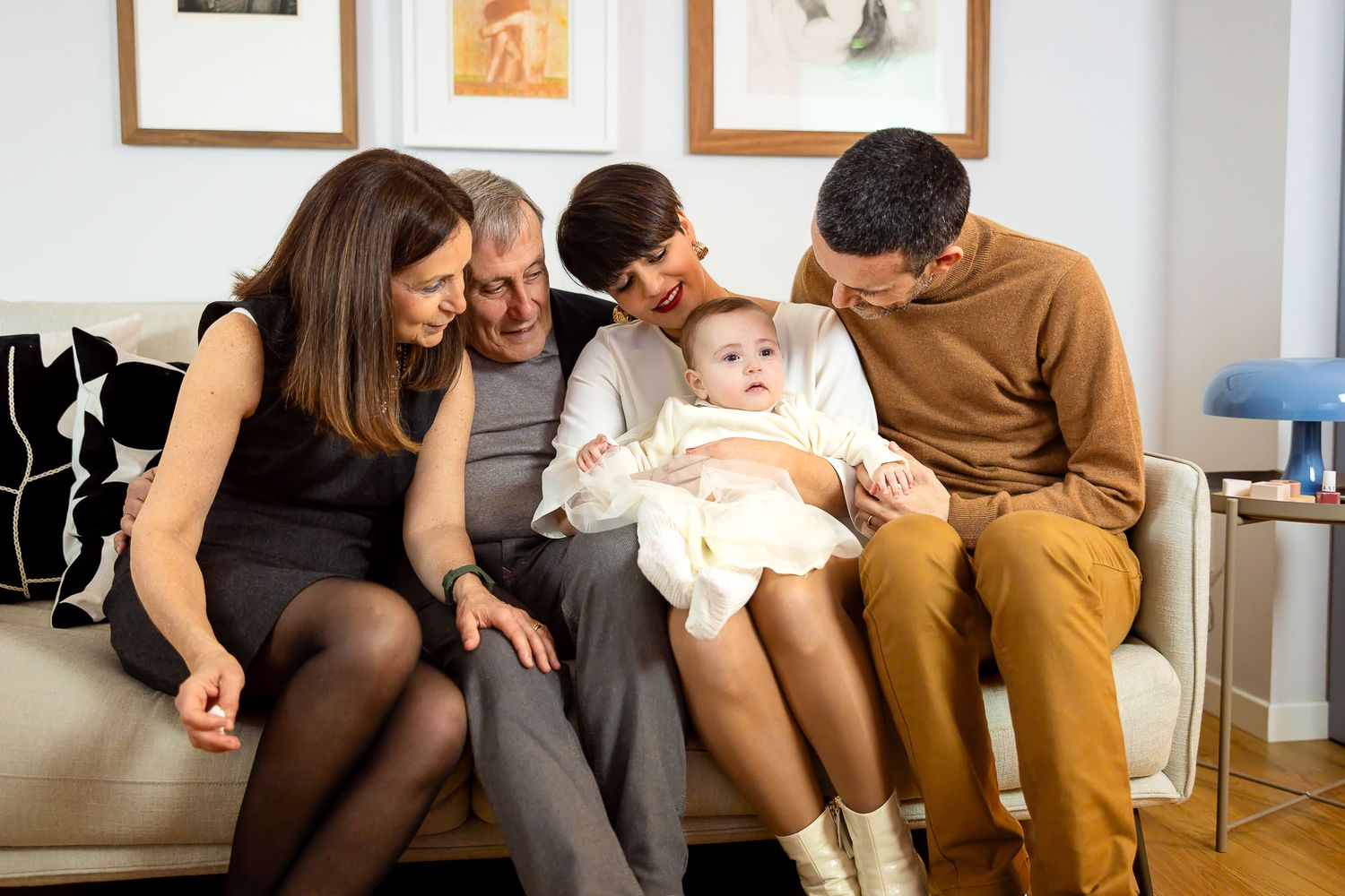 Happy multigenerational family sitting on sofa, smiling at adorable baby in white outfit during indoor gathering.