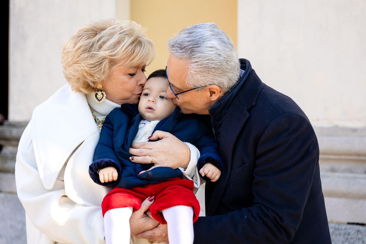 Grandparents kissing cheeks of baby dressed in blue and red outfit outdoors.